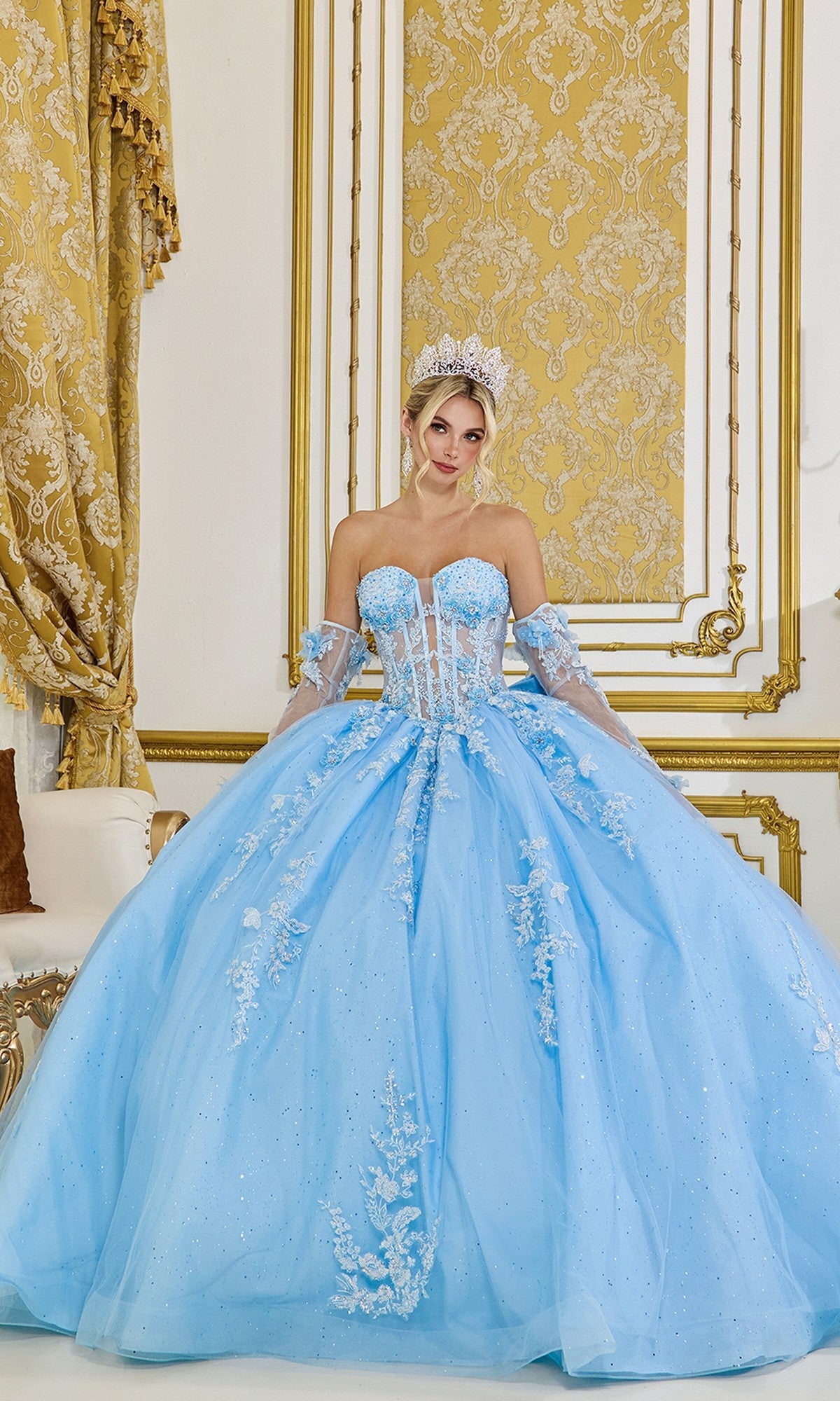 A woman stands in an ornate gold and white room wearing the Quinceanera Dress 5081 By Dancing Queen in Bahama Blue, featuring beaded lace applique, matching gloves, and a tiara.